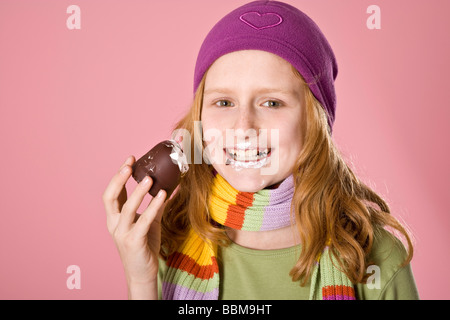 Fille aux cheveux roux portant un bonnet violet en face d'une toile rose, mordre dans une guimauve au chocolat Banque D'Images