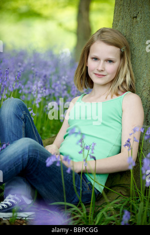 Jeune fille assise contre un arbre dans un bois plein de bluebells Banque D'Images