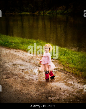 Petite fille dans un tutu rose et bottes rose sautant dans les flaques d'eau à côté d'un lac. Banque D'Images