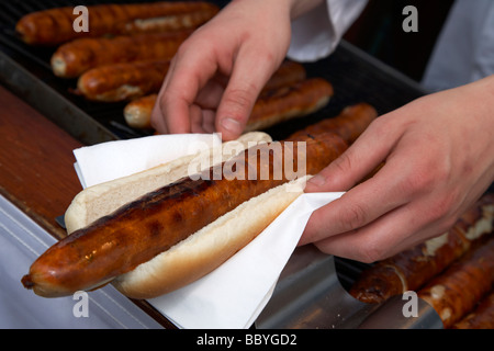 La préparation du vendeur à bratwurst pain bap sur serviette avec les saucisses allemandes sur le gril en vente sur un marché en plein air au Royaume-Uni Banque D'Images