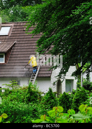 Homme portant un imperméable jaune debout sur une échelle de toit nettoyage avec un tuyau d'eau haute pression Banque D'Images