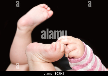 Close up horizontale d'un jeune bébé holding sur ses petits pieds avec sa main sur un fond noir Banque D'Images