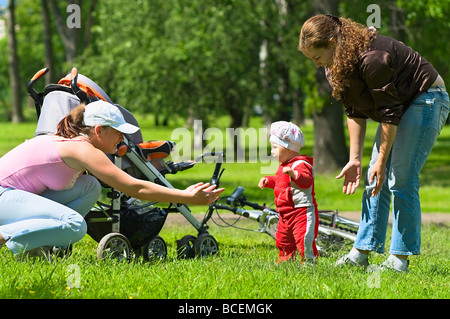 Deux femmes aider tout-petit à faire les premiers pas dans le parc. L'herbe verte et le feuillage autour d'elles. C'est un été. Poussette et vélo Banque D'Images