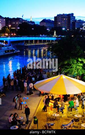 Donaukanal Riverside Bar et vie nocturne au crépuscule Vienne Autriche | VIENNE, Autriche — les foules se rassemblent dans un bar extérieur le long du Donaukanal (canal du Danube) au crépuscule alors qu’un catamaran Twin City Liner, qui dessert Bratislava, passe sur l’eau. Le canal est un bras régulé du Danube séparé de la rivière principale par des écluses, et pendant les mois les plus chauds, ses remblais deviennent l'un des quartiers de vie nocturne en plein air les plus populaires de Vienne, bordé de bars pop-up et de rues multiples Banque D'Images