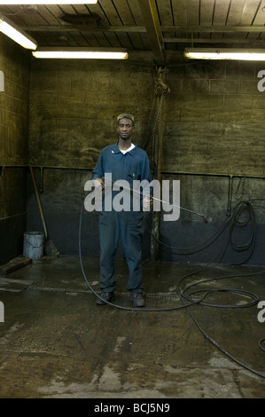 African American male in blue coveralls bretelles holding baguette de nettoyage vapeur appareil debout dans sale travail bay Banque D'Images