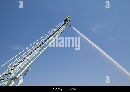 Pompier POMPIER sur haut de grande échelle avec jet d'eau de pulvérisation firehose against sky Banque D'Images