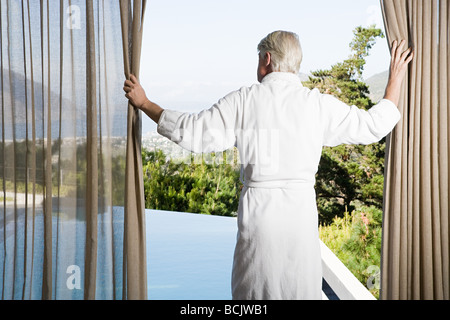Middle aged man wearing bathrobe tirant les rideaux en face de la piscine Banque D'Images