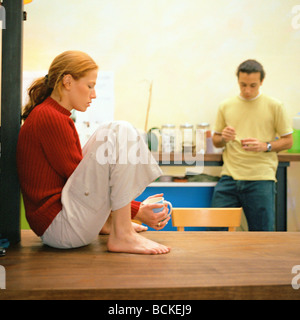 Jeune femme assise sur une table de cuisine, homme debout avec tasse Banque D'Images