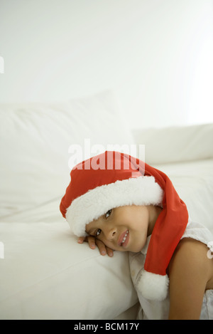 Girl leaning head sur canapé, wearing santa hat, smiling at camera Banque D'Images