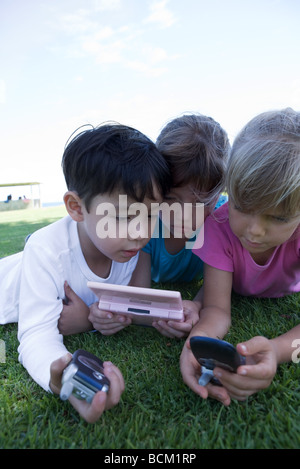 Trois enfants allongés sur l'herbe, jouer avec des jeux vidéo, maintenant des téléphones cellulaires, close-up Banque D'Images