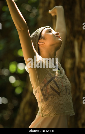 Young woman standing outdoors with arms raised, looking up, side view Banque D'Images