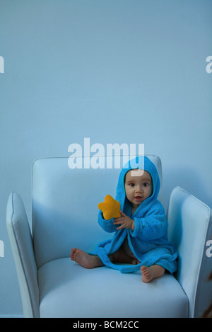 Baby Boy wearing hooded peignoir assis dans un fauteuil, tenant doudou, looking at camera Banque D'Images