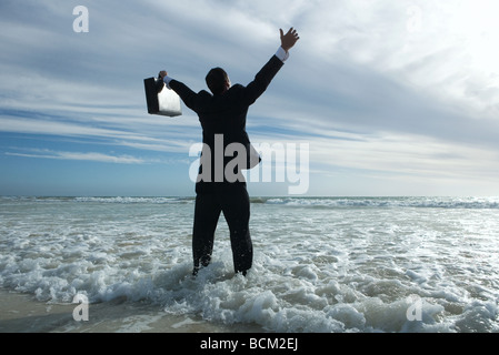 Businessman standing in surf at beach, bras levés, vue arrière Banque D'Images