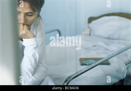 Femme assise sur le lit d'hôpital, regardant vers le bas avec la main sous son menton, cropped view Banque D'Images