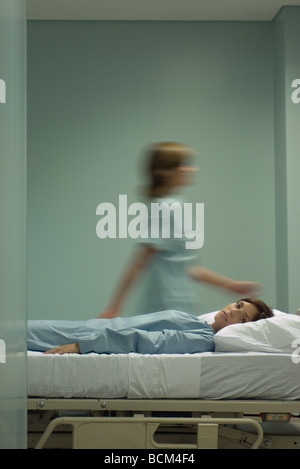 Woman lying on hospital bed, looking at camera, infirmière en marche en arrière-plan Banque D'Images