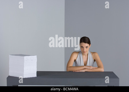 Femme assise à la table à côté d'une grande pile de papier, les bras croisés, looking at camera Banque D'Images