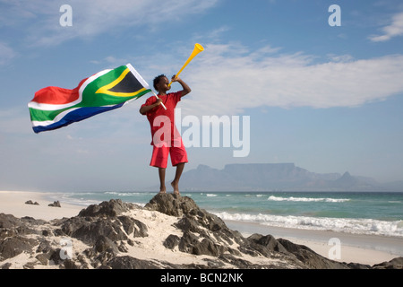 Jeune garçon soufflant sur vuvuzela, en maintenant drapeau sud-africain sur la plage, à la montagne de la table en arrière-plan, Blouberg Beach Banque D'Images