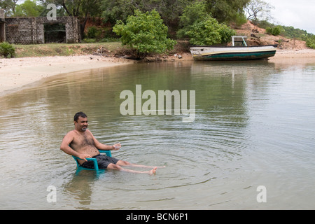 L'homme se détendre avec un verre sur une chaise dans l'eau jusqu'aux genoux à la mer les pêcheurs à Ceylan, Club Clappenburg Bay, Trincomalee, Sri Lanka. Banque D'Images