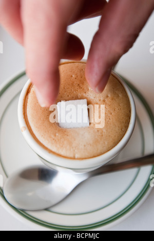 Vue aérienne d'une main féminine droping un cube de sucre dans la mousse d'un café expresso chaud boire dans une tasse avec une cuillère en métal Banque D'Images