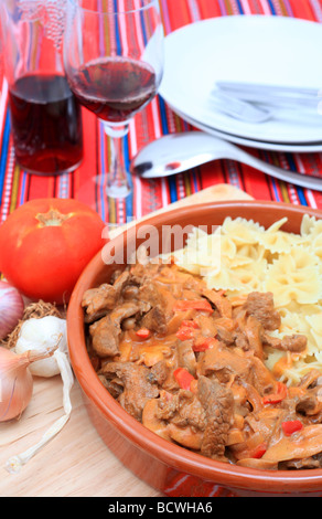 Un saladier en terre cuite avec bœuf stroganoff et farfalle sur une table avec des assiettes couverts et ingrédients culinaires vin Banque D'Images