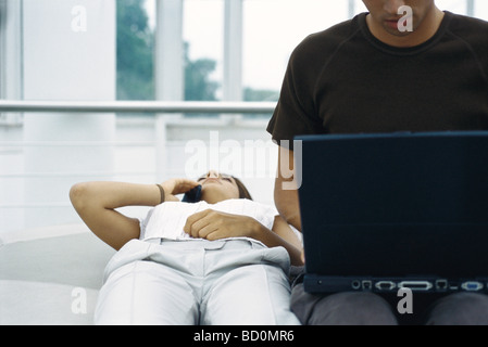 Jeune femme allongé sur le dos à l'aide d'un téléphone cellulaire, à l'aide de l'homme à côté de son ordinateur portable Banque D'Images