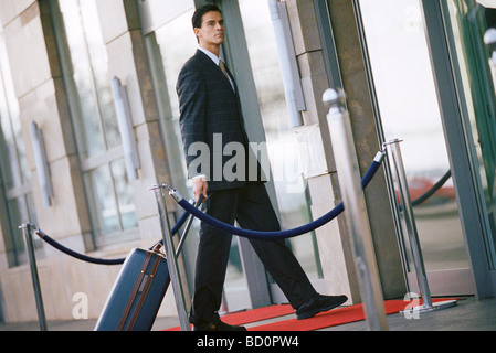 Businessman walking vers l'entrée du bâtiment, pulling suitcase derrière lui Banque D'Images