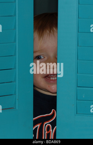 Un petit garçon rit comme il joue peek a boo derrière l'obturateur bleu windows. Banque D'Images