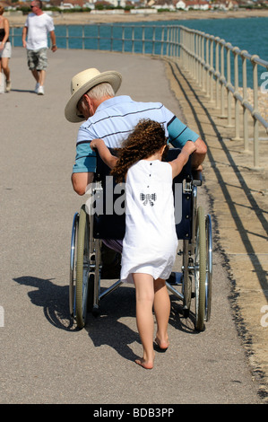 Petite fille poussant un adulte utilisateur de fauteuil roulant le long de la prom de Littlehampton England UK Banque D'Images