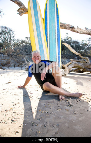 Homme d'âge moyen en wetsuit assis sur la plage en face de planches Banque D'Images