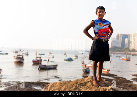 Un jeune garçon portant un tank top se dresse au sommet d'une pile de roche en face d'une baie pleine de bateaux de pêche. Banque D'Images