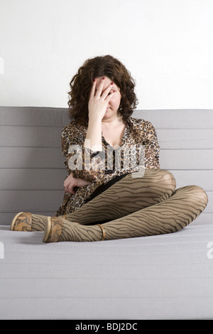 Young attractive woman chemisier léopard fermer les yeux par des mains assis sur un canapé avec les jambes dans des chaussures. Problème de femme. Bac blanc Banque D'Images