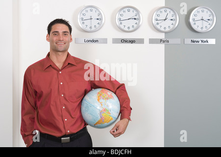 Businessman Holding Globe debout par Clocks Showing fuseaux internationaux Banque D'Images