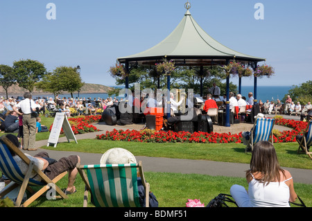 Vacanciers touristes assis dans des chaises longues à l'écoute d'un groupe de cuivres jouant dans le kiosque d'été Crescent Gardens Filey North Yorkshire Angleterre GB Banque D'Images