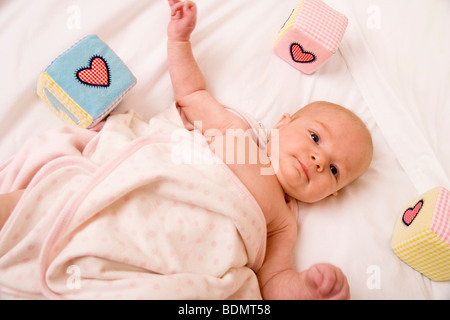 Petite fille de deux mois enveloppée dans une couverture douce, allongée sur un lit entourée de ses jouets colorés. Banque D'Images
