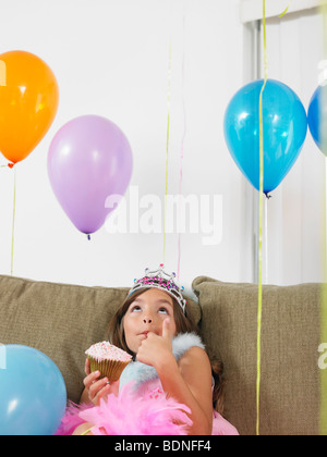 Jeune fille (7-9) sitting on sofa eating cupcake, looking up at balloons Banque D'Images