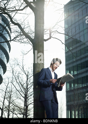 Jeune homme en litige en vertu de l'article arbre à l'extérieur de bâtiment de bureaux, reading newspaper Banque D'Images