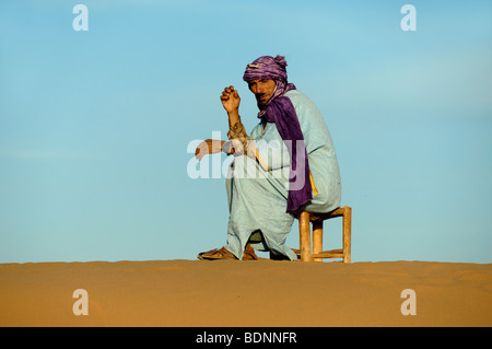 Homme berbère ou tribu portant des robes bleues traditionnelles, des vêtements, des vêtements ethniques ou une robe ethnique est assis sur un tabouret dans le désert du Sahara, près de Zagora, Maroc Banque D'Images