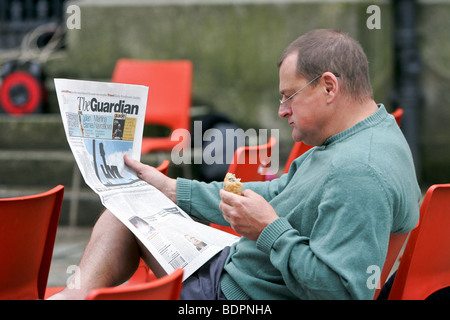 L'homme portant des lunettes s'asseoir et lire le journal The Guardian, tout en mangeant Banque D'Images