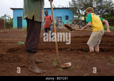 Un jeune garçon aux cheveux rouge blanc creuse une fondation dans le sol rwandais rouge supervisé par un homme. Banque D'Images