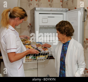 Femme âgée à l'aide d'un déambulateur en cuisine avec un soignant pour les services sociaux d'apprentissage pack les repas prêts dans son congélateur Banque D'Images