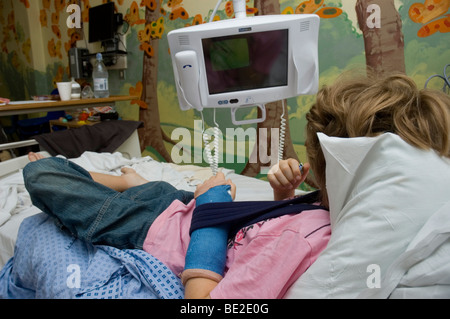 Un garçon avec un bras cassé se trouve sur un lit en regardant la télé en attente de traitement dans un hôpital du NHS Banque D'Images