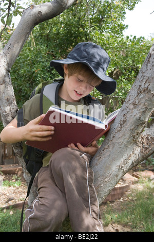 Sept ans la lecture dans un pommier, portant chapeau de soleil et un sac à dos Banque D'Images