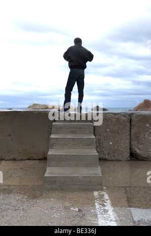 Un homme de grande taille est au-dessus de quelques marches de pierre sur un mur à la mer à l'horizon Banque D'Images