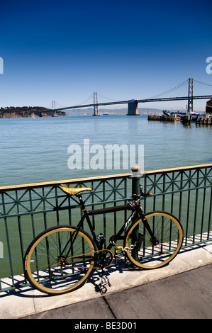 San Francisco Oakland Bay Bridge Bicycle Waterfront San Francisco Californie | SAN FRANCISCO, Californie — Un vélo à train fixe repose contre une rampe de front de mer, le San Francisco-Oakland Bay Bridge enjambant la baie derrière. Ouvert en 1936, le pont suspendu jumelé relie San Francisco à Oakland via l'île de Yerba Buena et était, à l'époque, le pont le plus long et le plus cher jamais construit. Banque D'Images