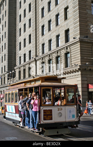 San Francisco Cable car numéro 19 avec Riders sur le côté Downtown San Francisco California USA // SAN FRANCISCO, California, United States — Cable car numéro 19, un véhicule classique du San Francisco Municipal Railway (muni), est vu sur sa route dans le centre de San Francisco. Les passagers roulent à la fois à l'intérieur et debout sur le côté du véhicule emblématique, un spectacle commun sur ces lignes historiques. Le panneau de la voiture indique qu'elle dessert les lignes populaires Powell & Market et Hyde & Beach Fisherman's Wharf, soulignant son rôle en tant qu'attraction touristique majeure. Le système de téléphérique de San Francisco, un National Historic Banque D'Images