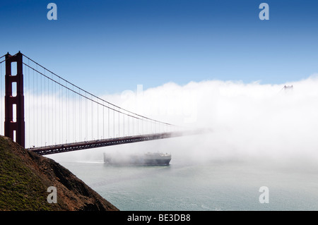 Golden Gate Bridge Ship émergeant du brouillard San Francisco Californie | SAN FRANCISCO, Californie — Un cargo émerge d'une rive de brouillard sous le Golden Gate Bridge, sa tour éloignée et sa route avalée par la couche marine que les habitants appellent « Karl the Fog ». Achevé en 1937, le pont suspendu s'étend sur 1,7 miles (2,7 km) à travers le détroit du Golden Gate, avec des tours s'élevant à 746 pieds (227 m) au-dessus de l'eau. L'acier orange international de la tour proche contraste fortement avec le ciel bleu clair au-dessus de la rive nuageuse. Banque D'Images