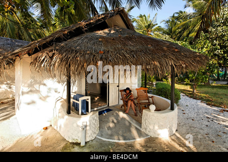 Femme avec chapeau assis sur une chaise en face d'un bungalow, porte ouverte, palmiers, les Maldives island, South Male Atoll, Maldives, Ac Banque D'Images