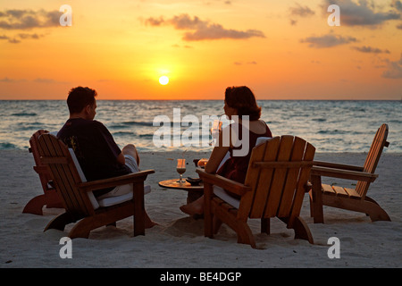 Couple assis sur des chaises à la plage avec des long drinks devant un coucher du soleil doré, Rihiveli, Island, Maldives, South Male Ato Banque D'Images