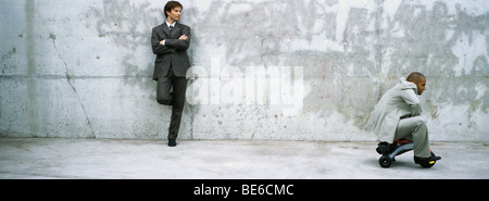 Businessman with arms folded leaning against wall ride associé à regarder l'enfant tricycle Banque D'Images