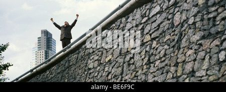 Businessman standing on wall with arms raised, gratte-ciel en arrière-plan Banque D'Images